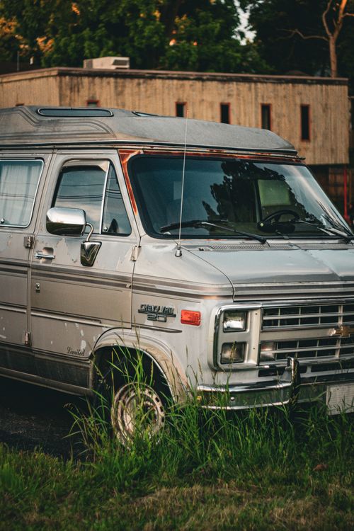 Free Classic silver van in tall grass with building backdrop under warm sunlight. Stock Photo