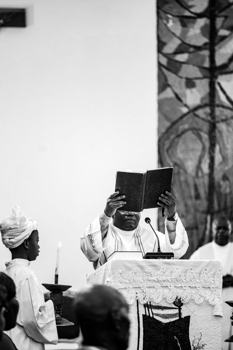Black and white photo of a priest holding a bible during a sermon in a Kinshasa church with a child assisting.