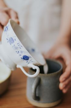Close-up of milk being poured from a flowery porcelain cup into a clay mug.