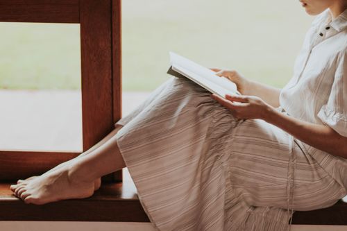 Free A woman sits barefoot on a windowsill, reading a book in natural light. Stock Photo