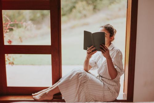 Free A woman in a white dress sitting by the window, reading a book in a serene setting. Stock Photo