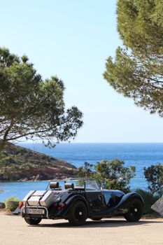 Classic vintage car parked by the scenic ocean coast amid trees, under a clear blue sky.