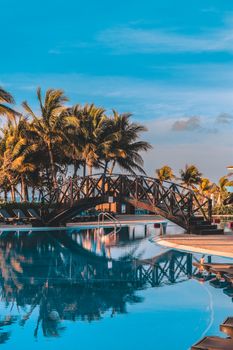 Stunning view of a resort's pool featuring a picturesque wooden footbridge amidst palm trees, creating a tropical getaway vibe.