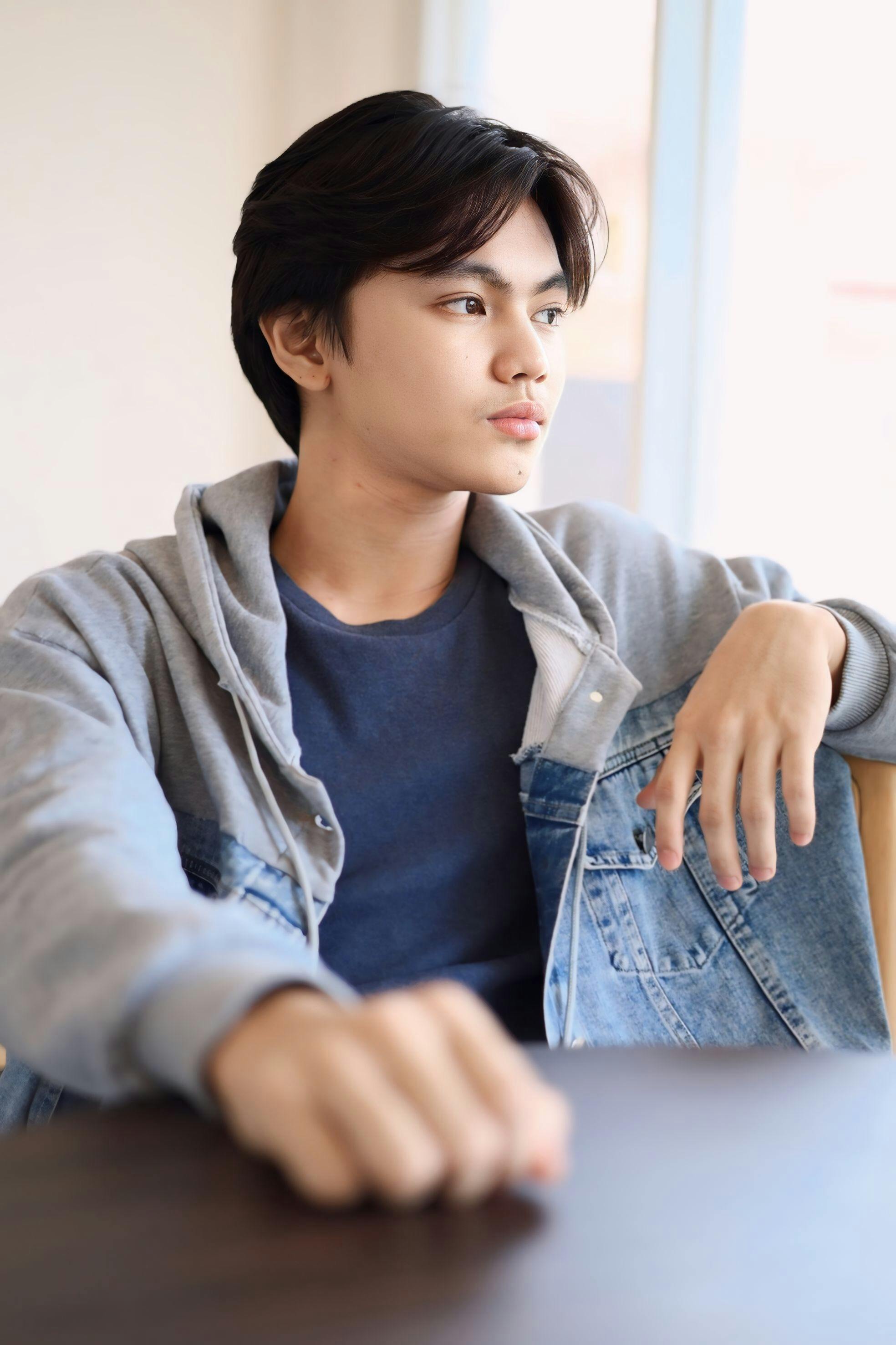Trendy teenager in casual denim jacket sitting indoors, exuding cool and relaxed vibes.