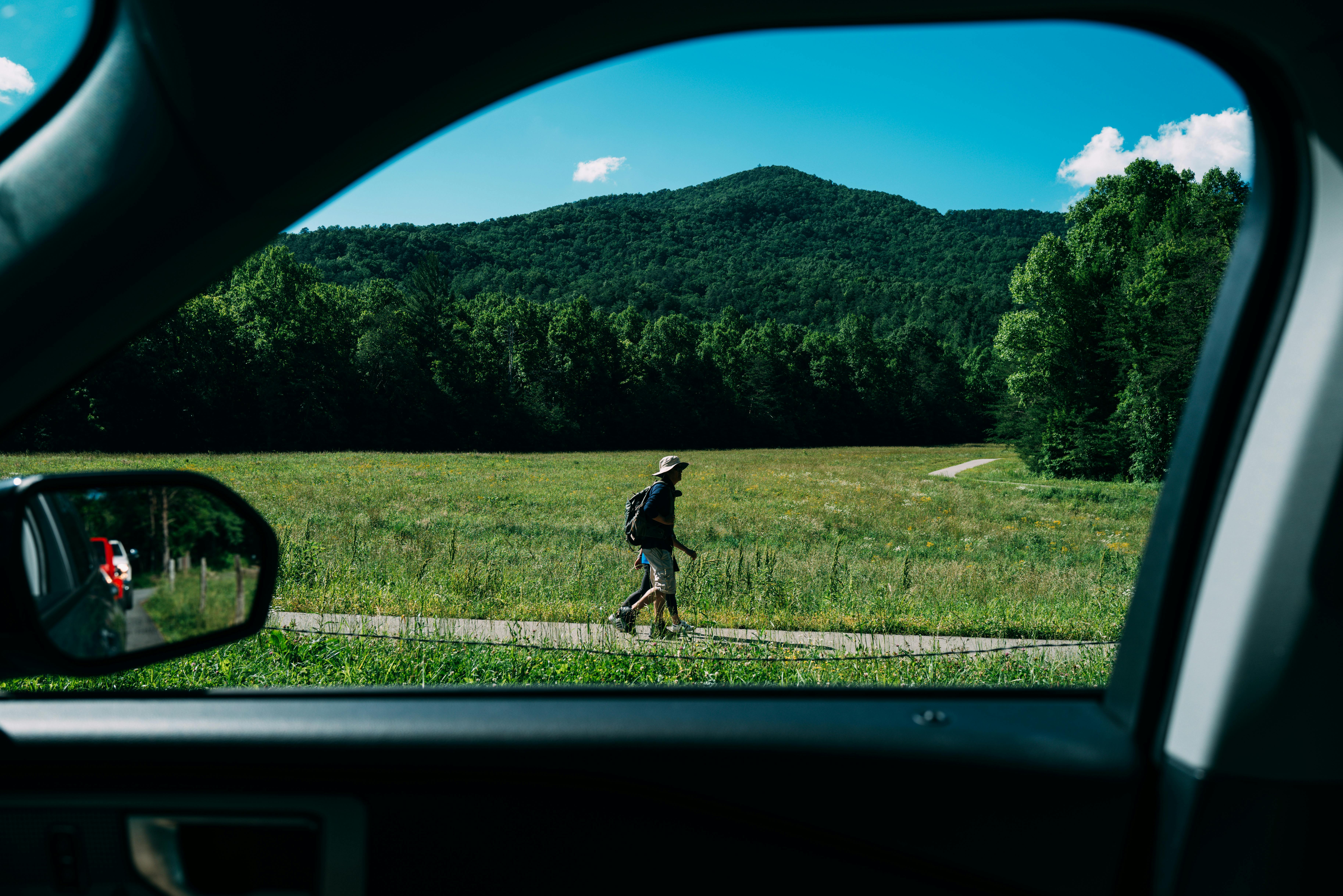 A man hikes through a grassy valley viewed from a car window, with mountains in the background.