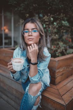 一个戴着眼镜、穿着牛仔夹克的时髦女人正在户外品尝咖啡