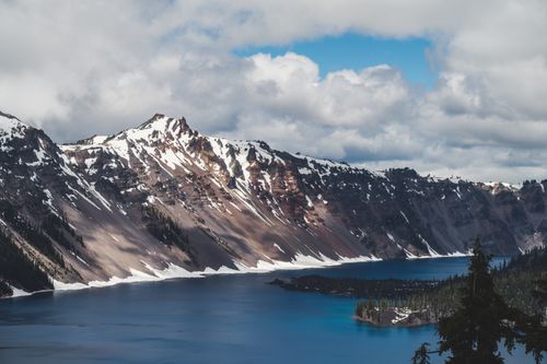 Free A stunning landscape of snow-capped mountains surrounding Crater Lake under a cloudy sky. Stock Photo