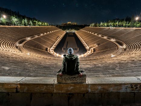 Man sitting at illuminated Panathenaic Stadium in Athens under a starry night sky.