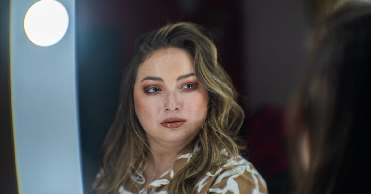 A fashionable woman with makeup looks at her reflection in a lit mirror, captured in a studio setting.