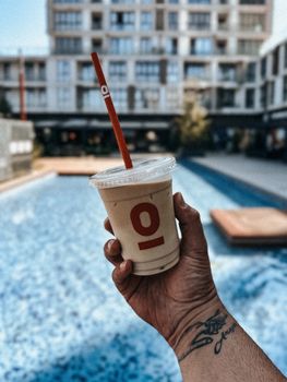 Hand holding an iced drink in a cup with a straw, poolside at a luxury resort during summer.