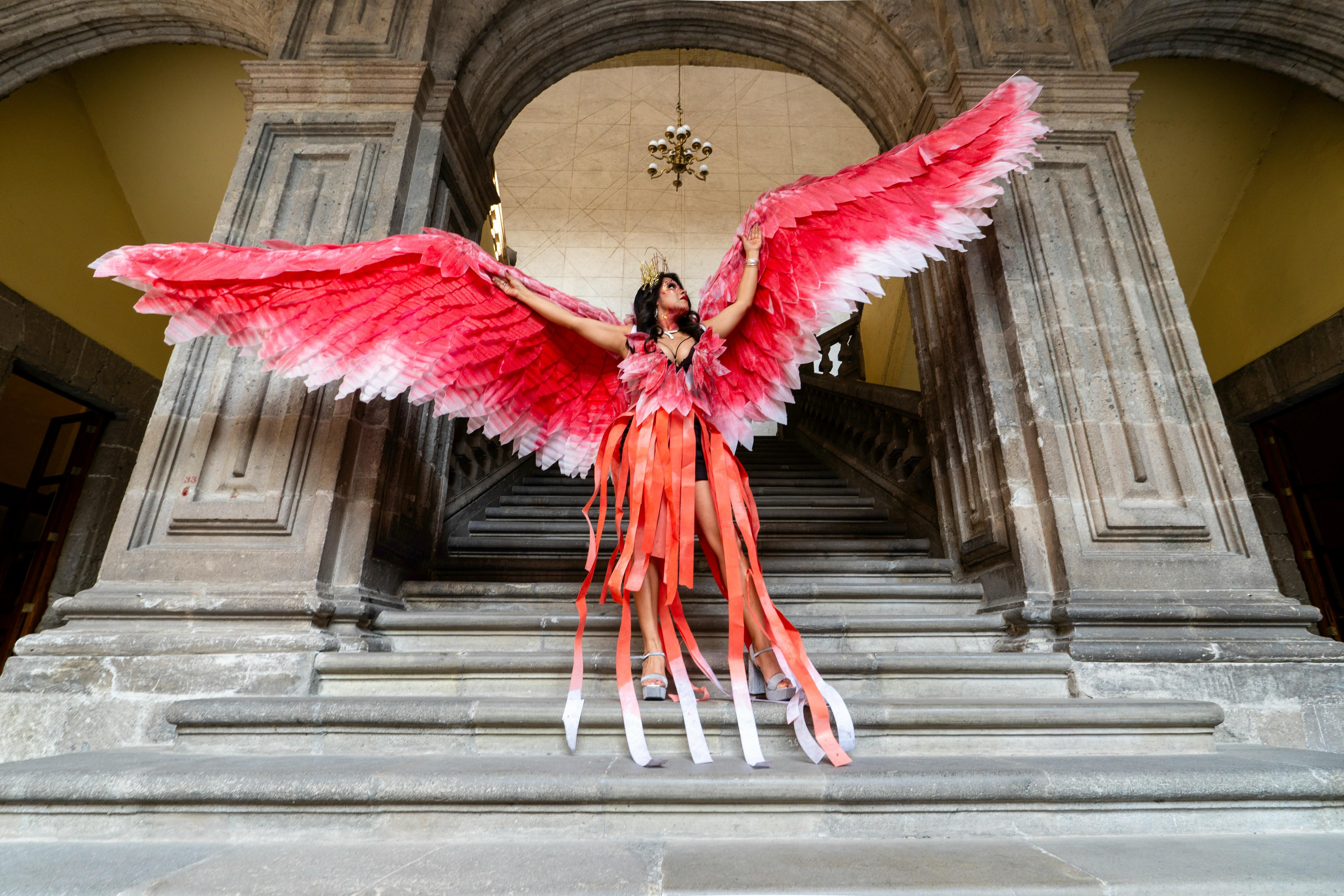 A woman poses in a vibrant red-winged costume on grand stone stairs in Mexico City.