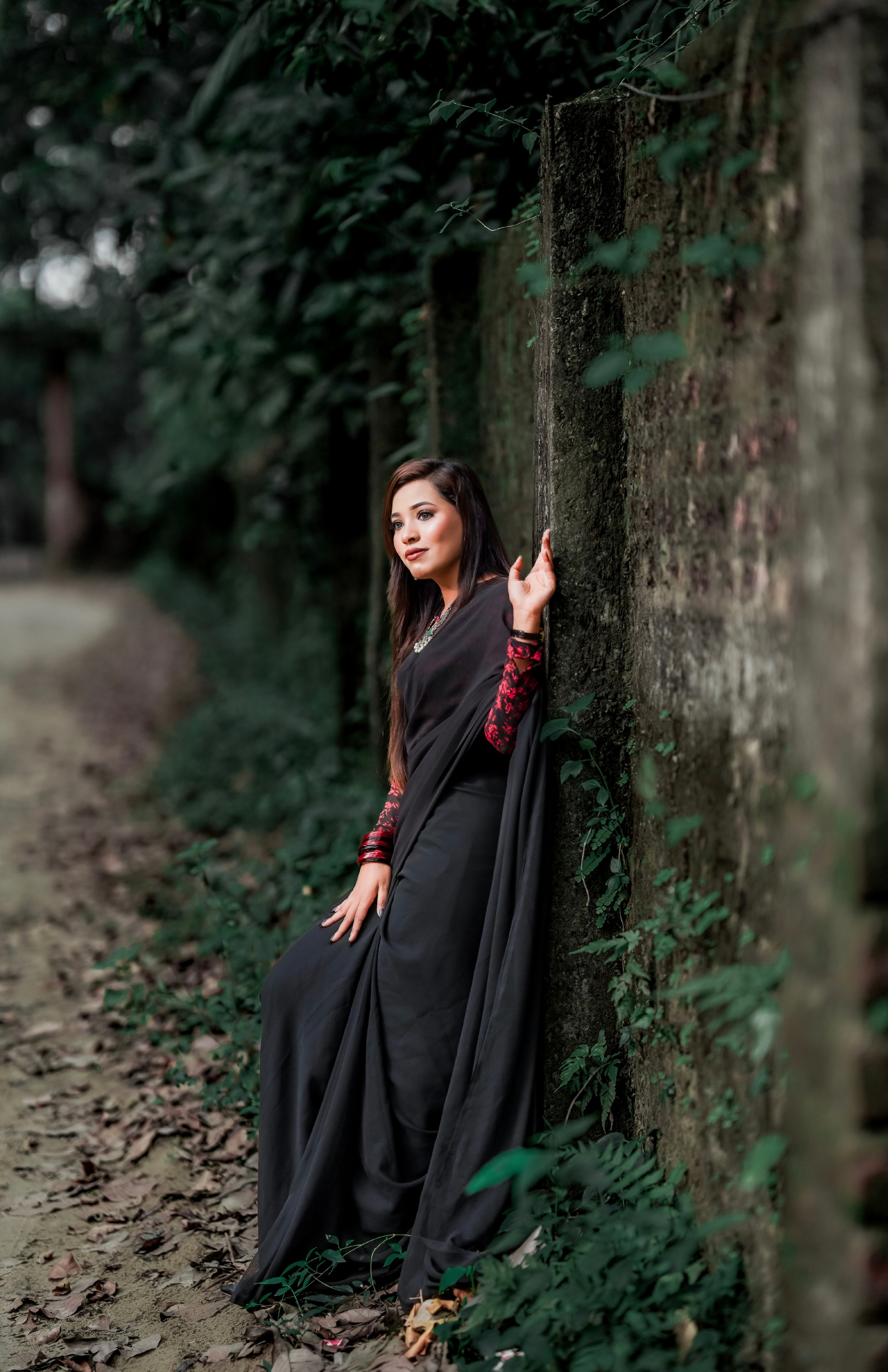 Fashionable woman in a black dress posing by an old wall in a verdant park, showcasing elegance.