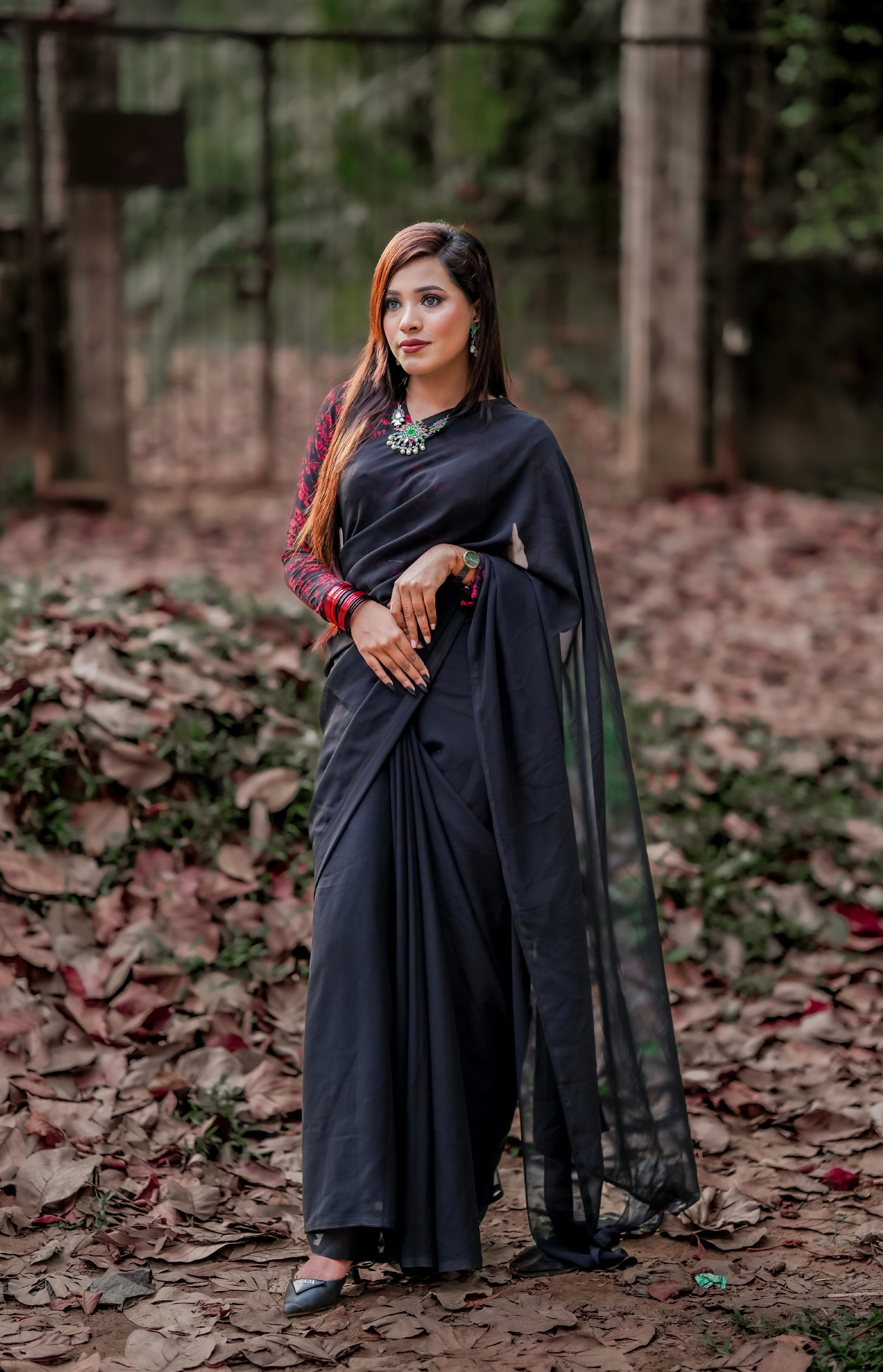A woman elegantly poses in a dark saree surrounded by fallen autumn leaves, exuding grace and style.