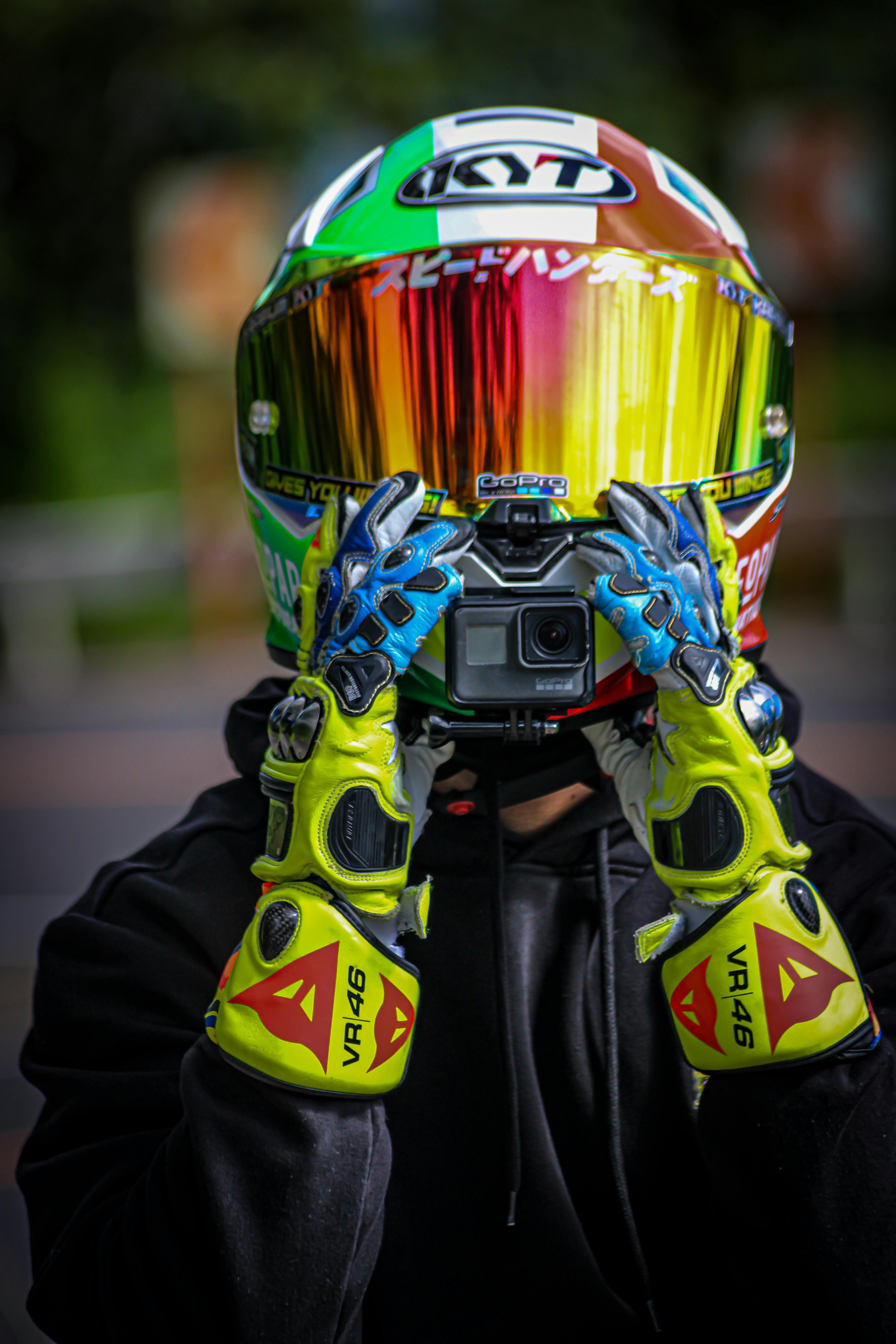 A motorcyclist in vibrant gear holding an action camera with reflective visor helmet.