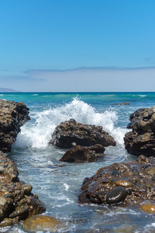 Free Vibrant ocean waves crash against eroded rocks on a sunny coastline with clear blue skies. Stock Photo