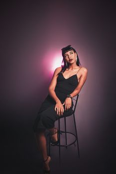 A stylish woman in a black dress posing confidently on a stool in a studio setting.