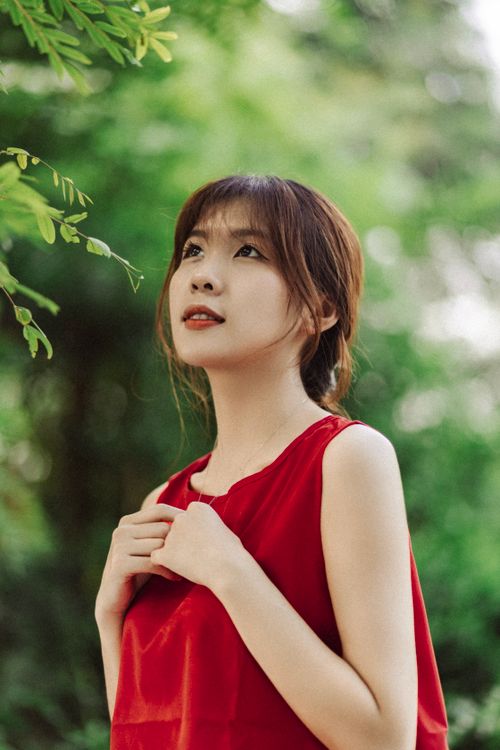 Free Portrait of a woman in a red blouse, looking upwards amidst lush green foliage. Stock Photo