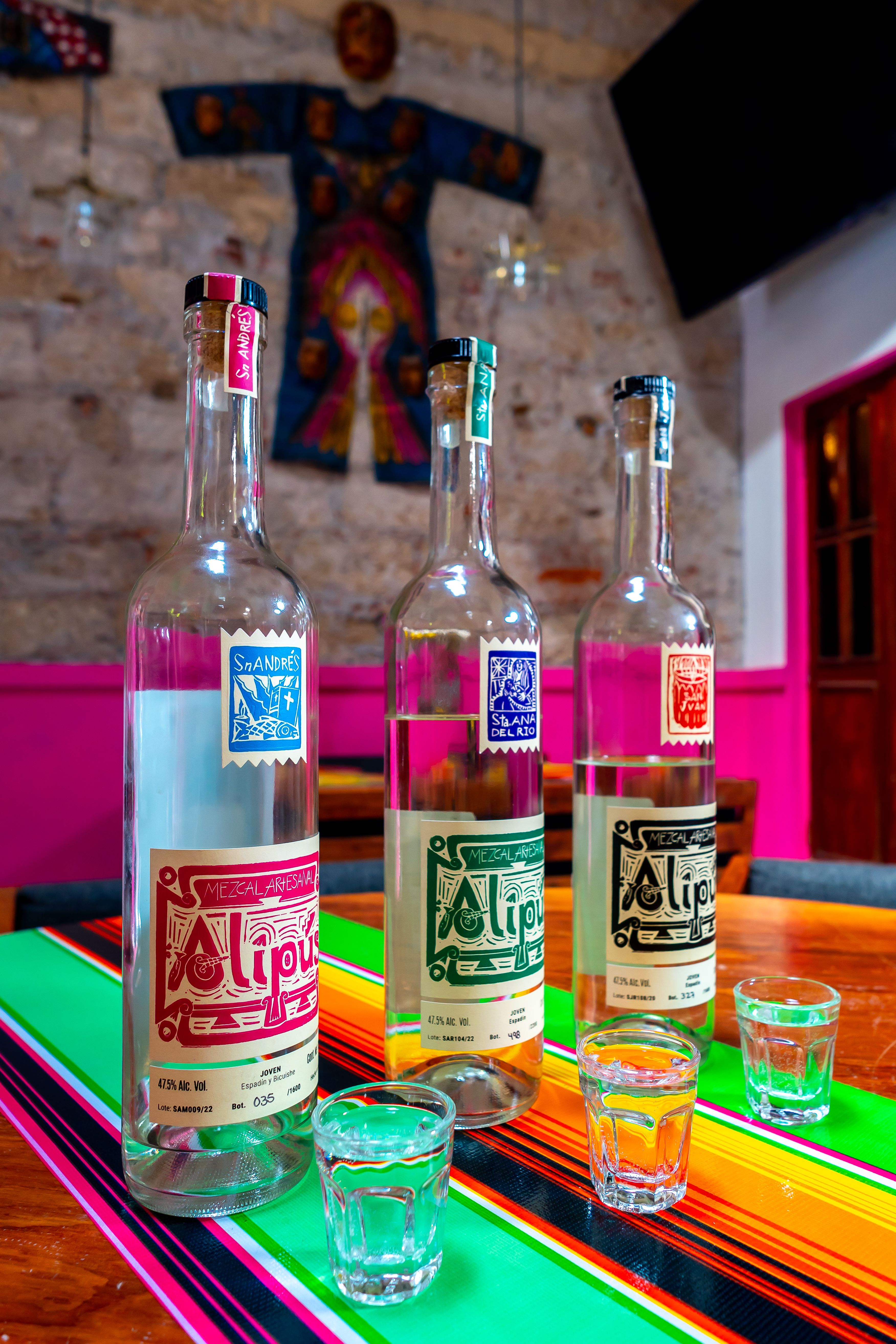Vibrant display of mezcal bottles and shot glasses on a table in a colorful Ciudad de México bar.