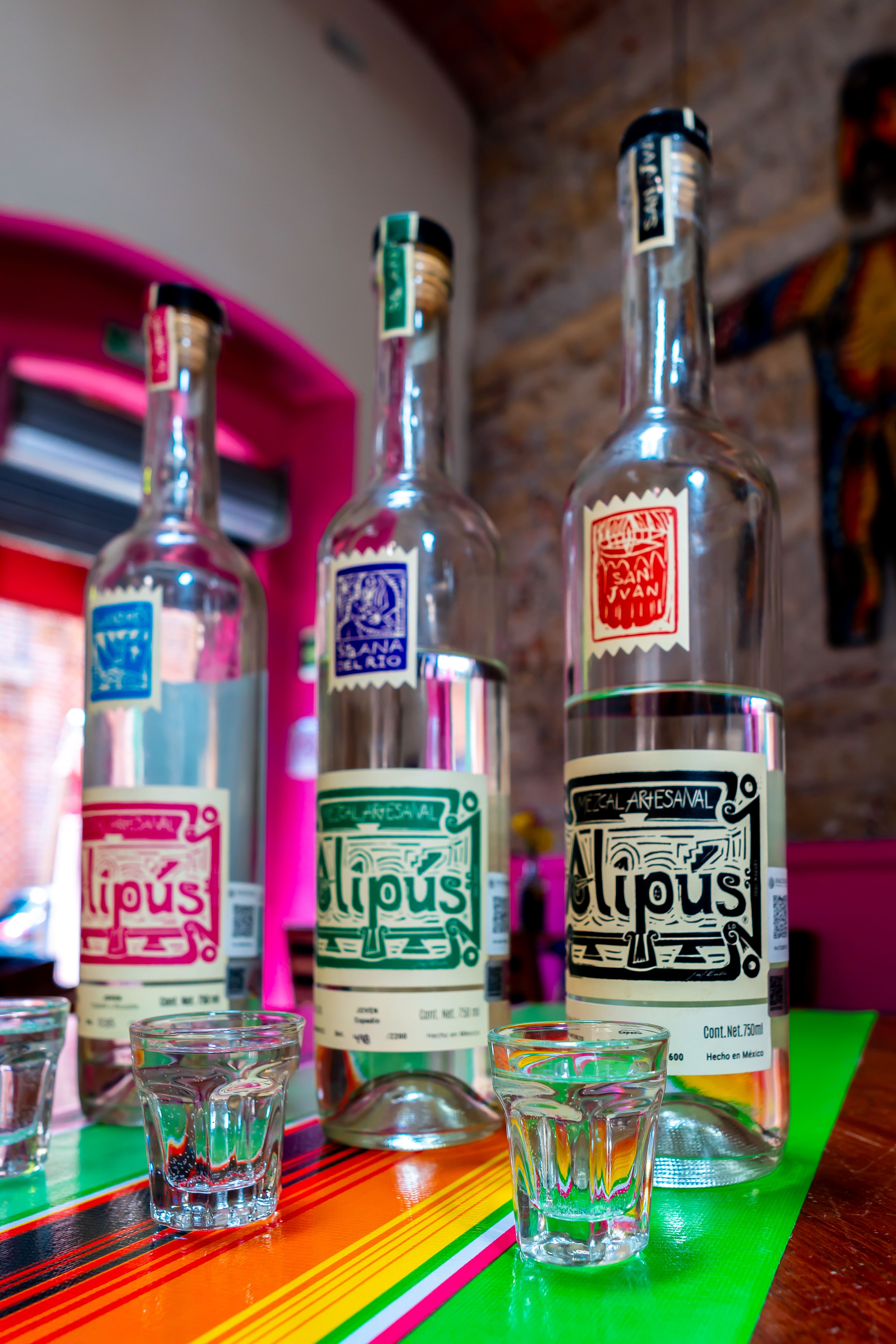 Three artisanal mezcal bottles with glasses on a colorful table in a Mexican bar.
