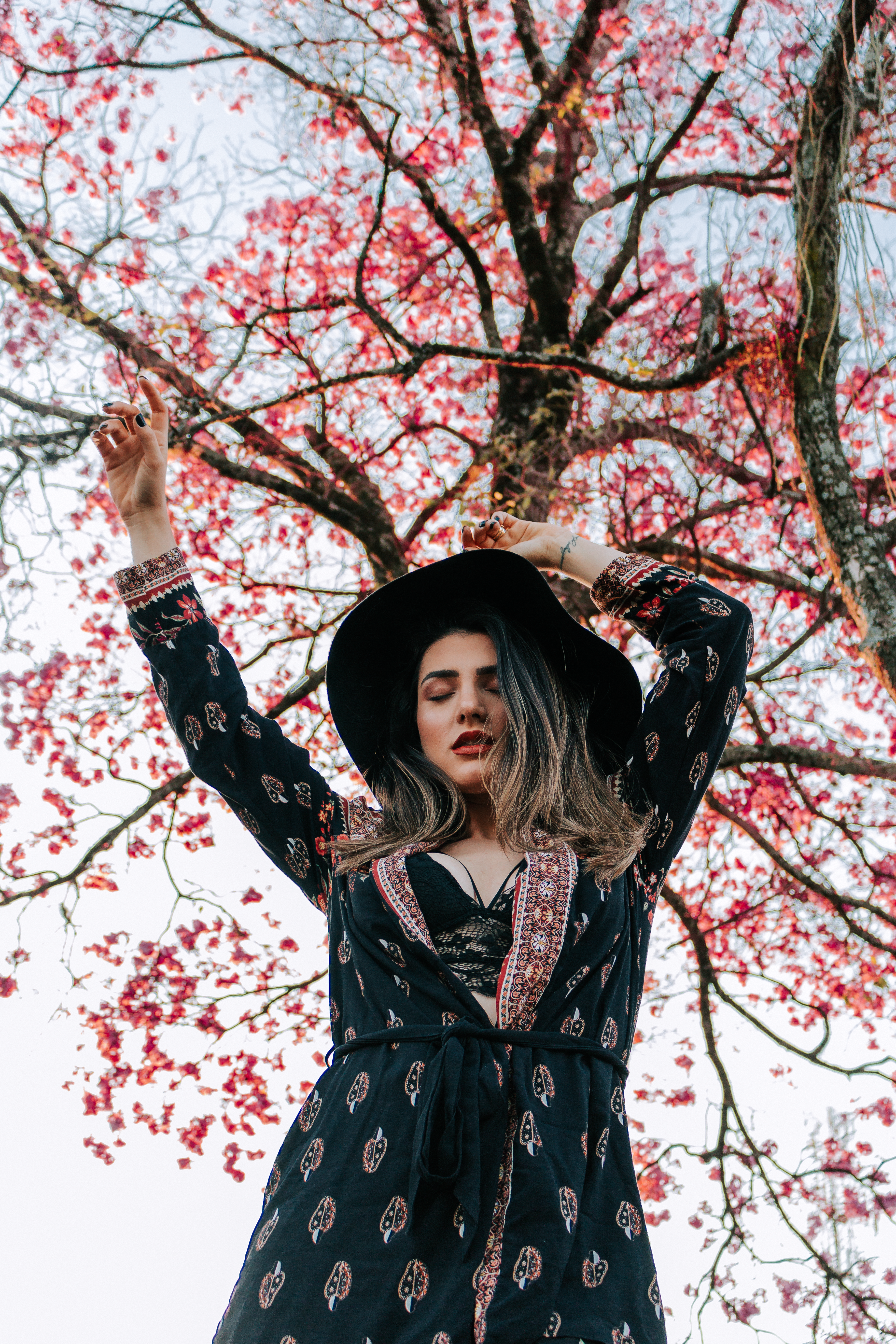 Fashionable woman in hat poses under a vibrant flowering tree in Medianeira, Brazil.
