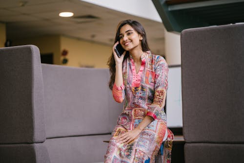 Free Photo of Smiling Woman in Floral Salwar Kameez Talking on Phone While Sitting on Edge of Wooden Table Stock Photo