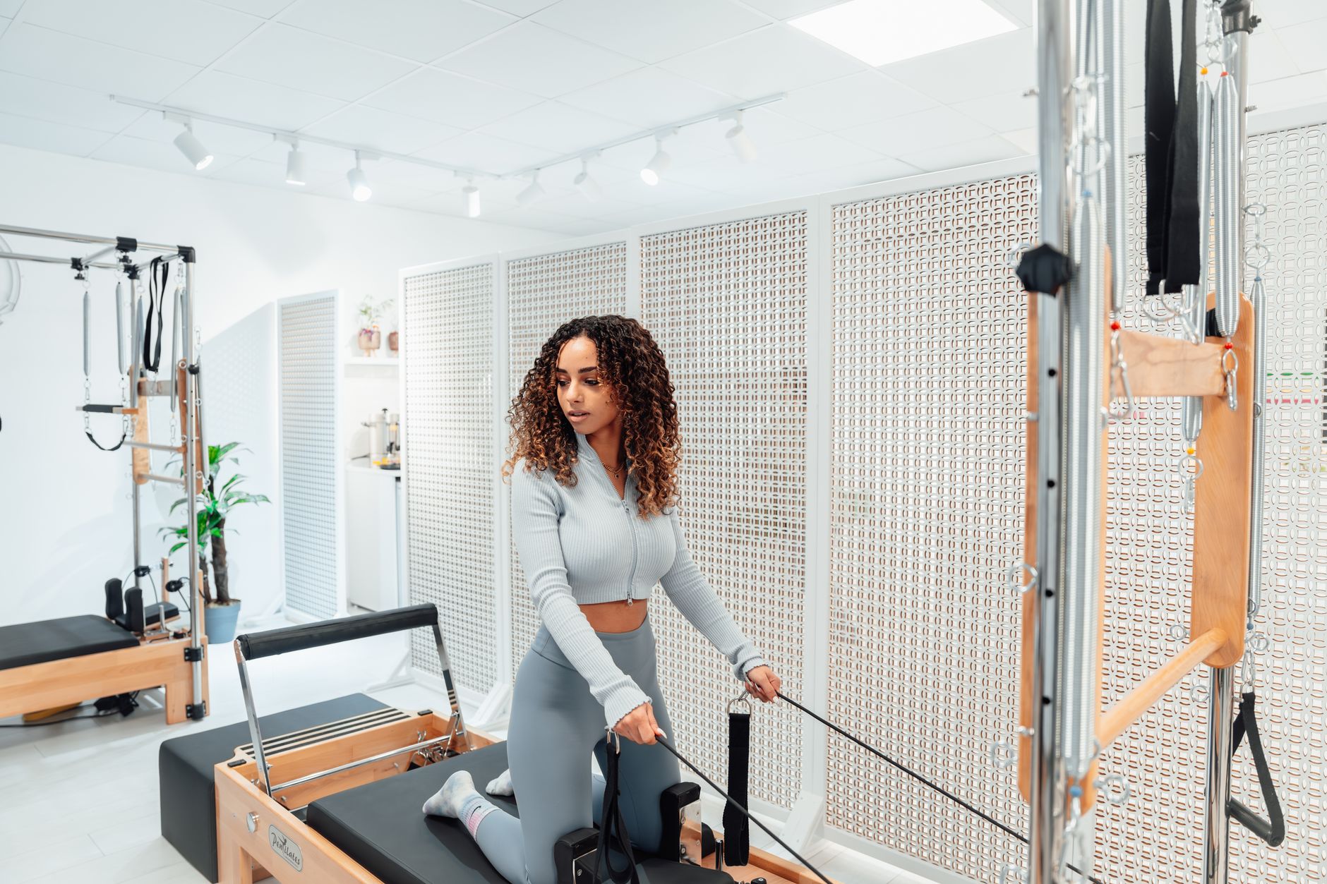 A woman in a gray tracksuit exercises on a Pilates tower in a bright, white room.