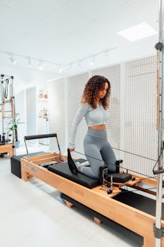 Woman in gray tracksuit exercises on Pilates tower in a bright white studio, showcasing fitness and concentration.