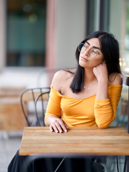 Photo of Women in Yellow Top Sitting by Table Posing with Her Eyes Closed