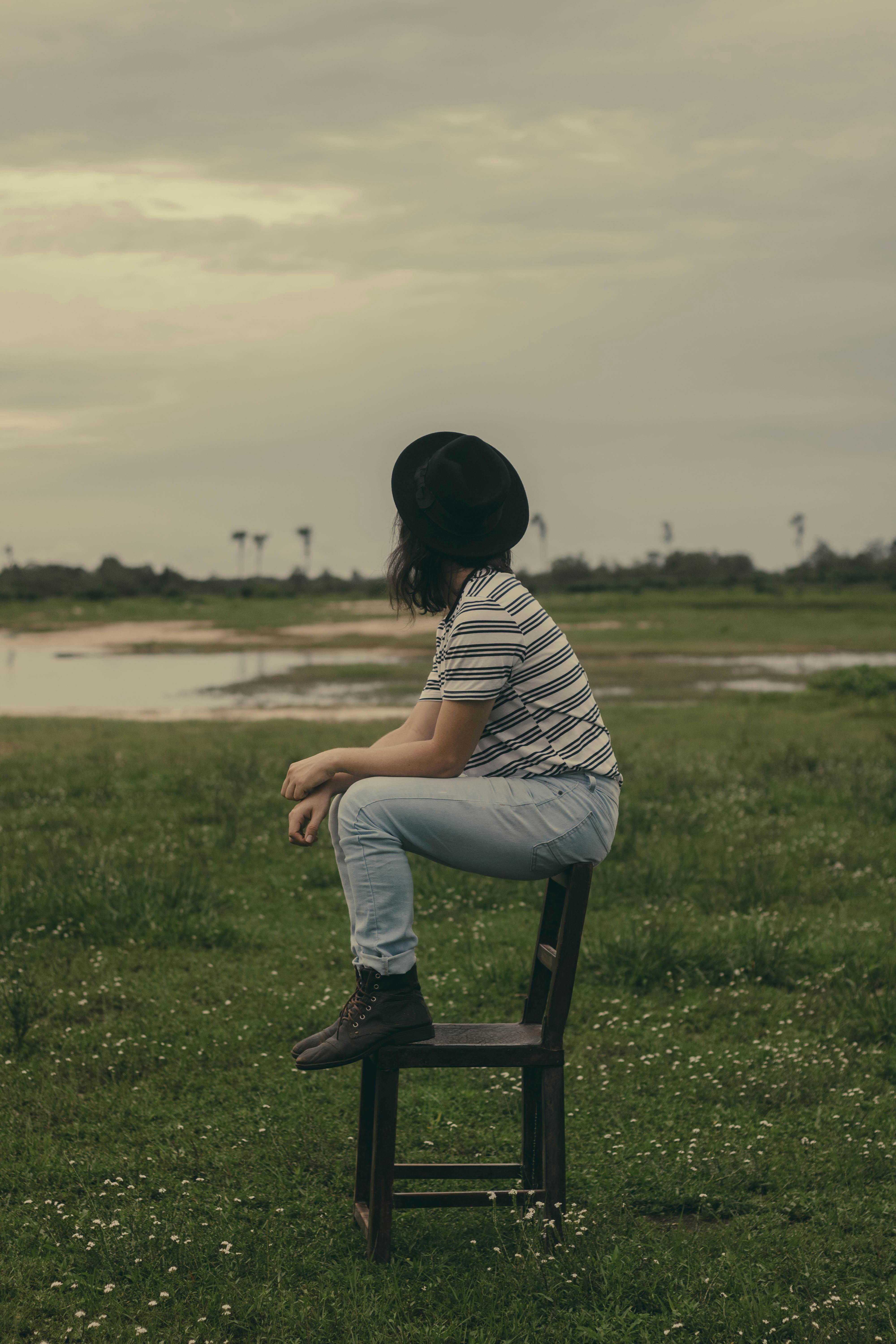 photo of woman sitting on brown wooden chair in the middle of a grass field looking away