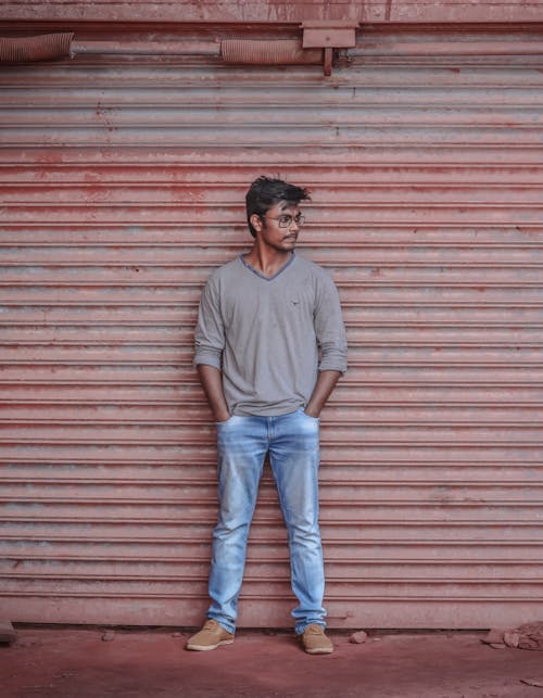Foto De Um Homem Em Uma Camiseta Cinza De Mangas Compridas E Calça Jeans Posando Em Frente à Porta De Enrolar Olhando Para Longe