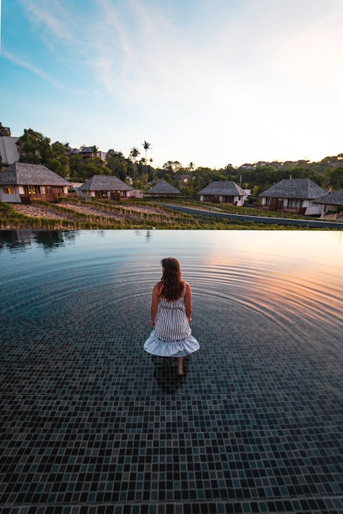 Back View Photo of Woman in White Dress Standing in the Middle of an Infinity Pool Looking at Resort Cottages