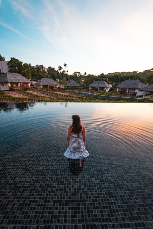 Free A woman in a white dress stands in an infinity pool at a Thai resort during sunset. Stock Photo