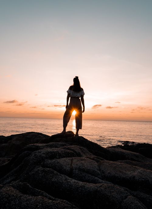 Free A lone woman stands on rocks, silhouetted against a beautiful sunrise by the sea in Thailand. Stock Photo