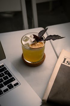 A citrus cocktail with ice and garnish on a modern office desk beside a laptop and newspaper.