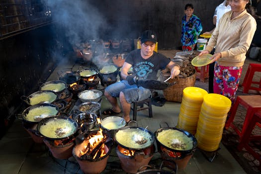 A street vendor cooking multiple Vietnamese pancakes over open flames in a bustling market setting.