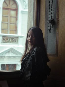 A young woman in a leather jacket smiling and standing by an urban window indoors.