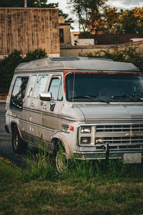 Free A vintage Chevrolet van parked outdoors, showing signs of rust and decay, surrounded by nature. Stock Photo