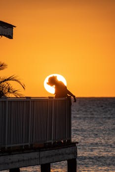 Silhouette of a woman leaning against a railing at sunset by the ocean, with warm yellow tones.