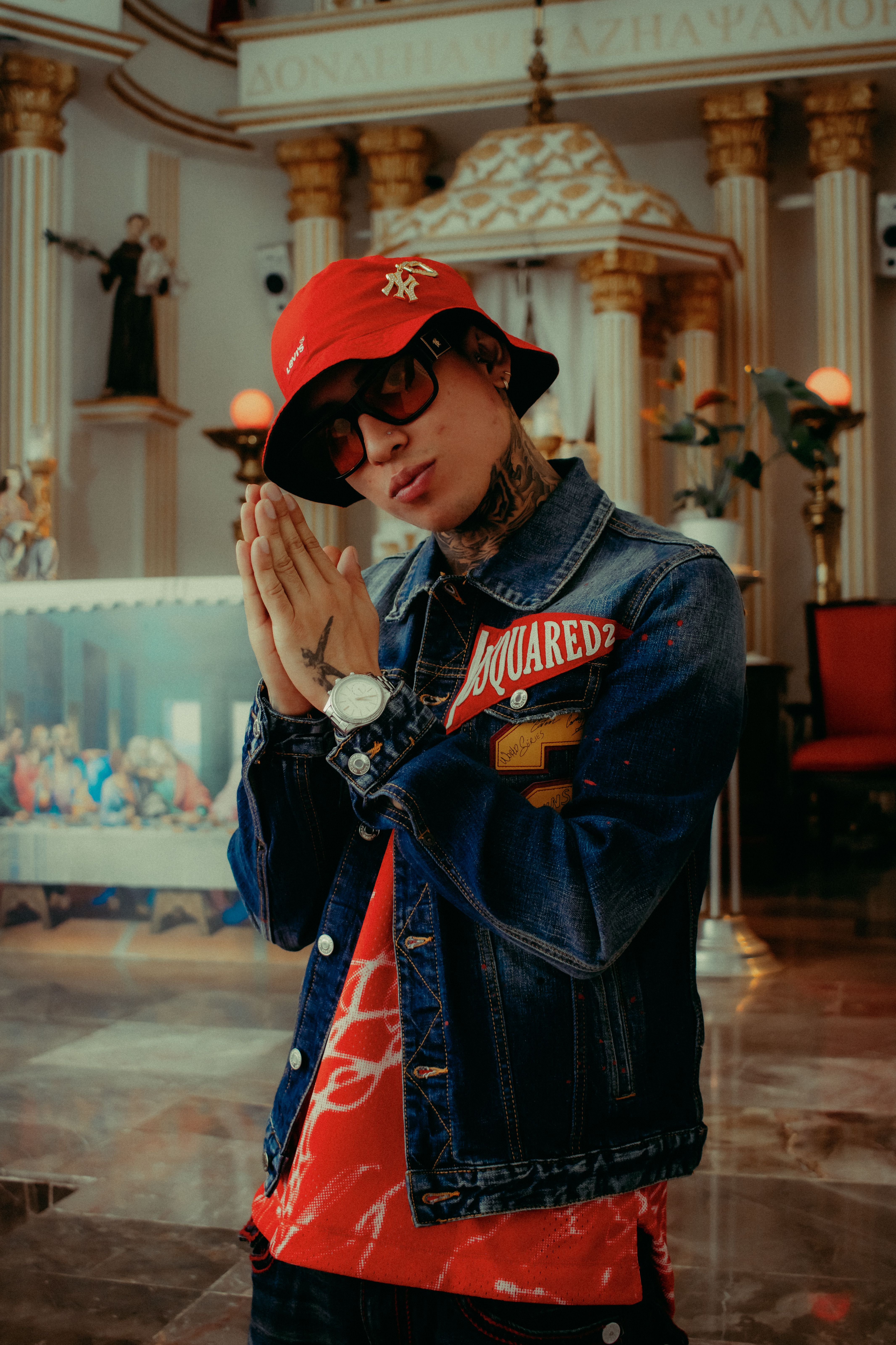 Fashionable young man in denim jacket and hat posing indoors with a prayer gesture.