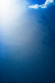 Serene aerial view of a calm blue ocean with a single cloud reflecting on the water.