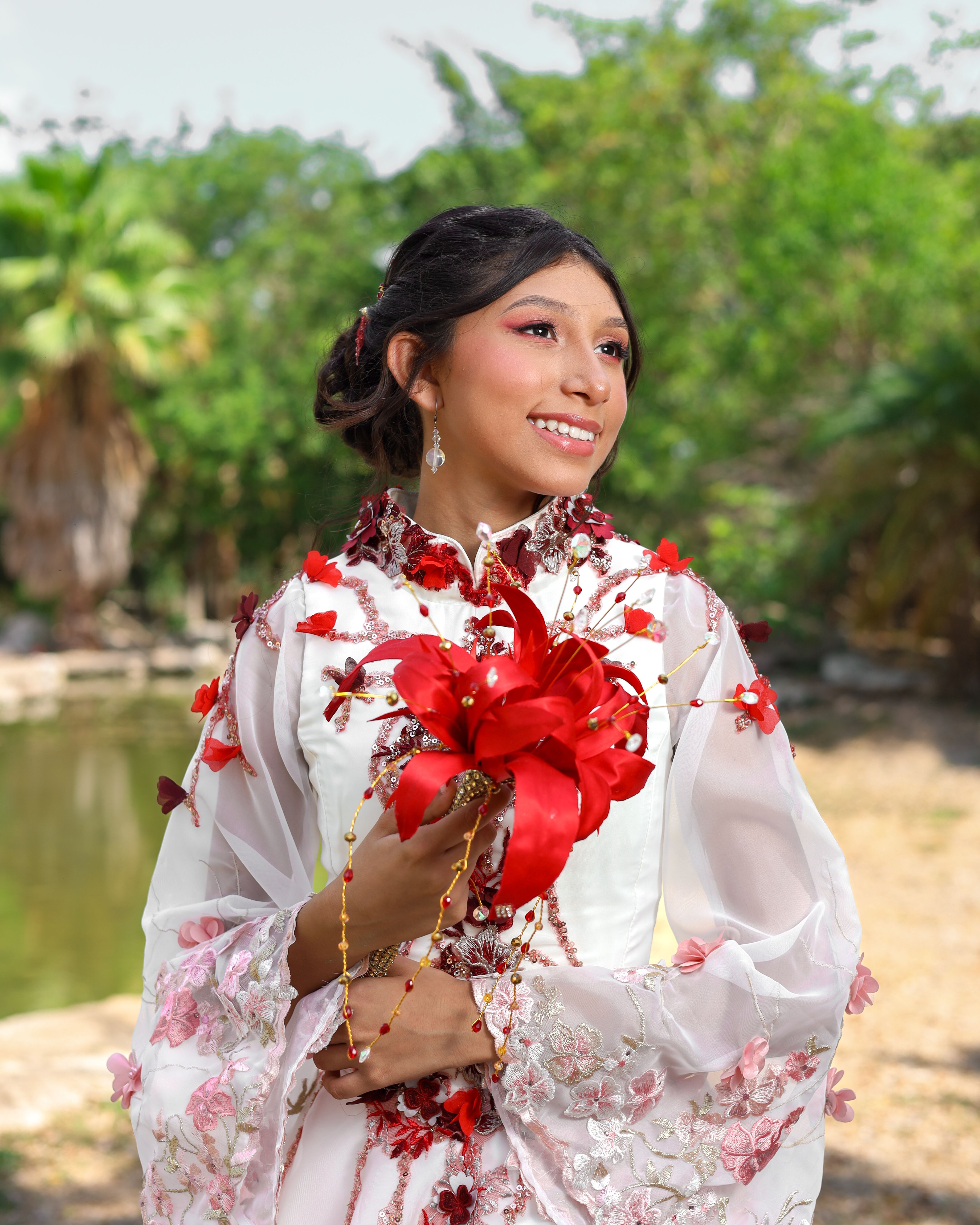 A young woman in a floral gown holding red flowers, standing outdoors in Mexico.