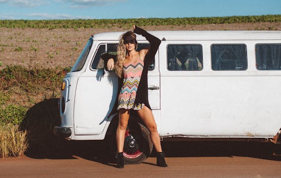 A woman poses against a vintage minivan on a sunny rural road, evoking a bohemian travel vibe.