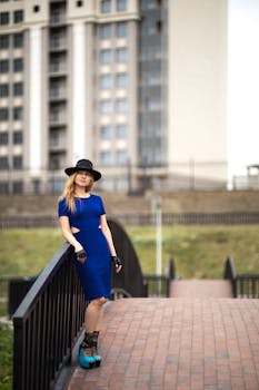 一位穿着蓝色连衣裙、戴着帽子的时尚女子，正倚靠在户外的桥栏杆上