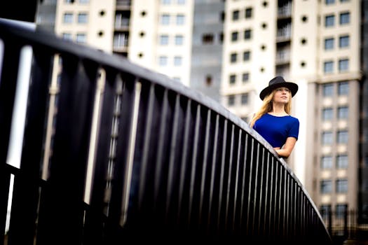 一位穿着蓝色连衣裙、戴着帽子的时尚女子在现代建筑背景的城市桥上摆姿势