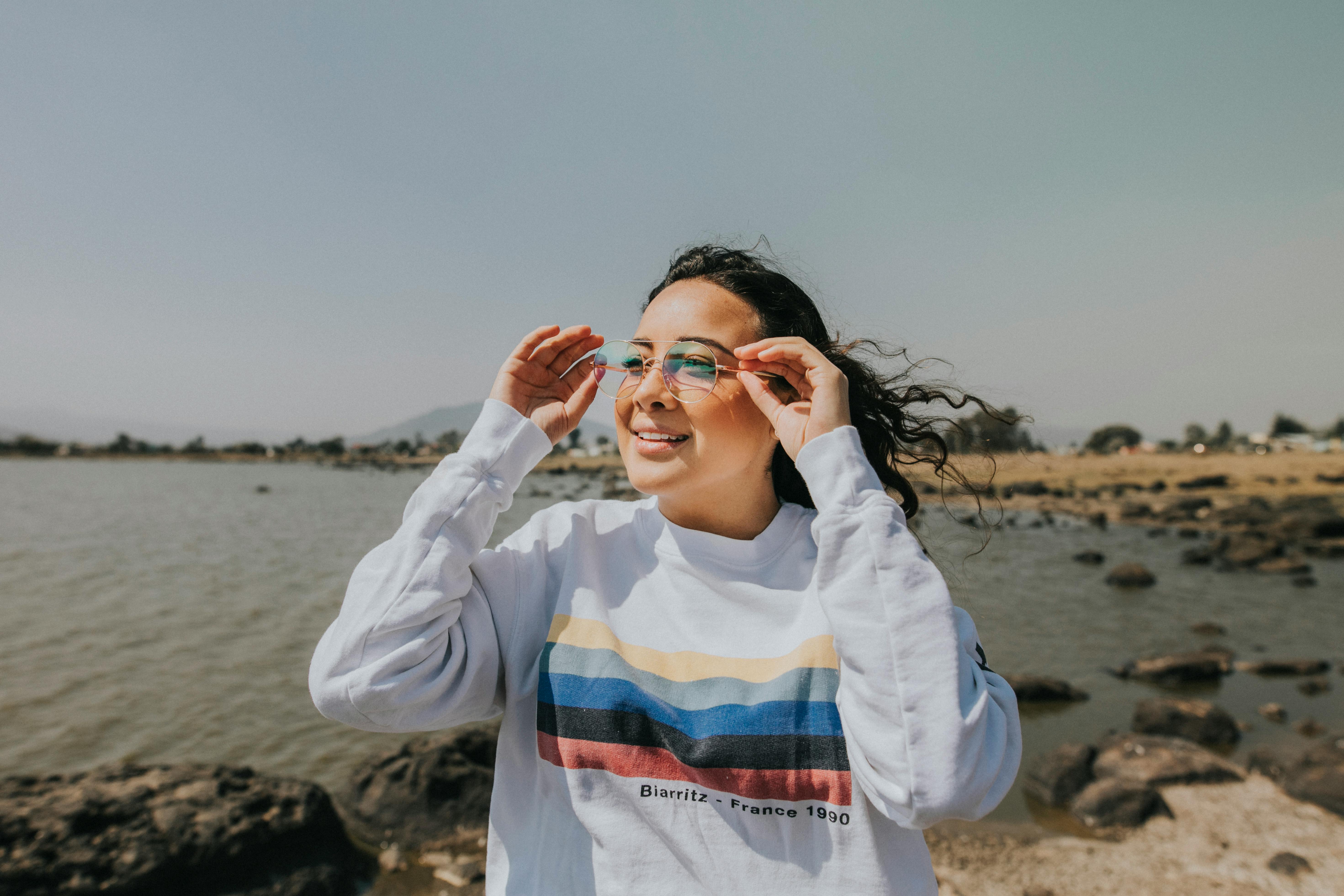 一名年轻女子在岩石海岸微笑着调整眼镜，享受阳光明媚的一天