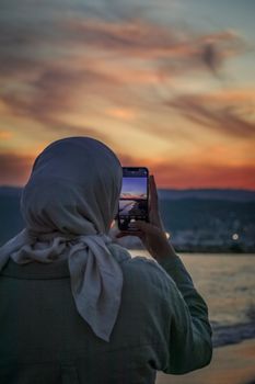 Woman in hijab photographing a stunning sunset by the seashore in Mersin, Türkiye.