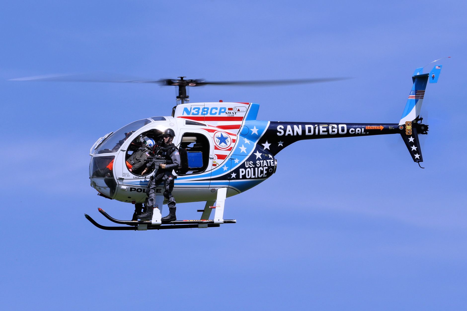 San Diego police helicopter with armed officer in flight against clear blue sky.