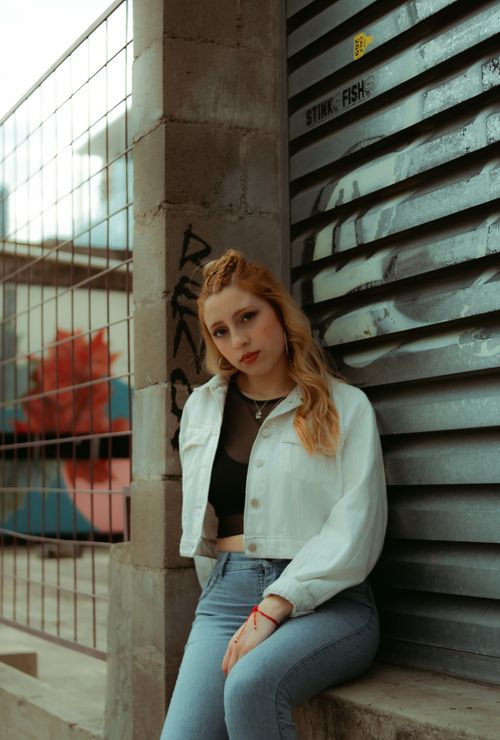 Free Young woman leaning against industrial backdrop, wearing casual denim attire in a modern urban setting. Stock Photo