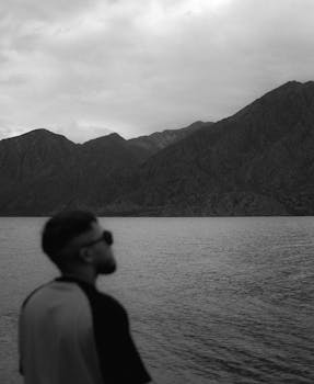 A serene black and white photo of a man by a lake with mountainous backdrop.