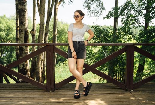 A woman in casual summer attire standing on a boardwalk in a sunlit forest area.